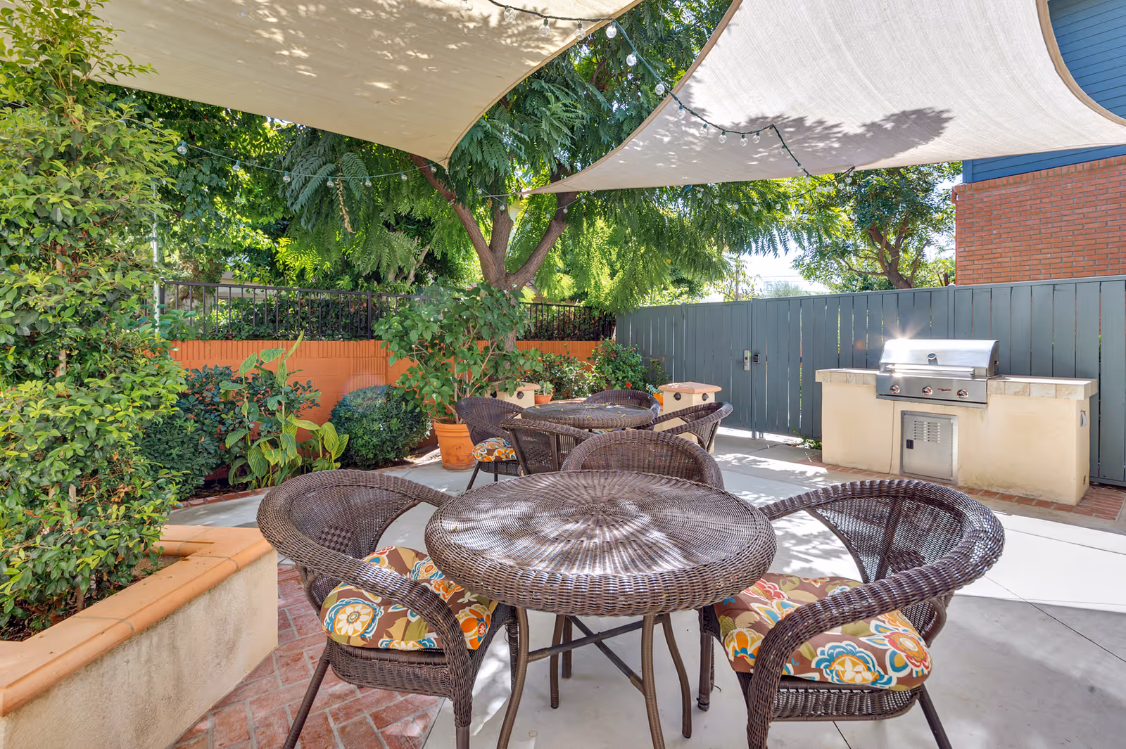 Outdoor patio area with wicker tables and chairs featuring colorful floral cushions. The space is shaded by large fabric canopies and surrounded by lush green plants and trees. A built-in stainless steel grill is visible against a gray wooden fence.
