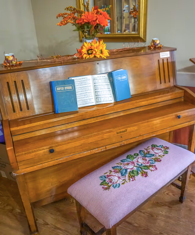 Wooden upright piano with open sheet music and hymnals, topped with autumn flowers and a mirror, and a floral-embroidered bench in front.