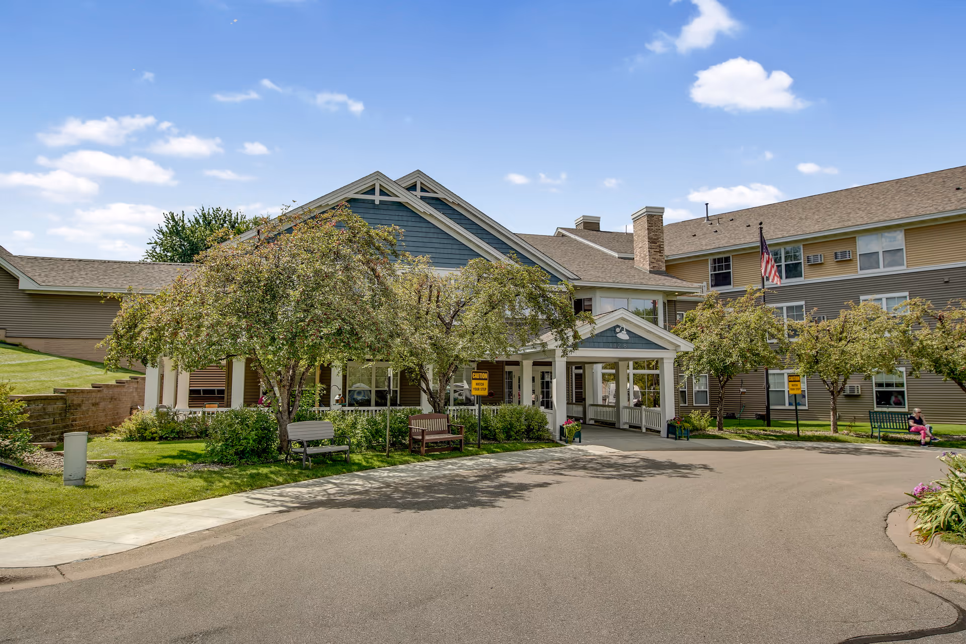 Front exterior view of Oak Ridge Assisted Living facility showing a large building with a covered entrance, trees, benches, and a clear blue sky with some clouds.