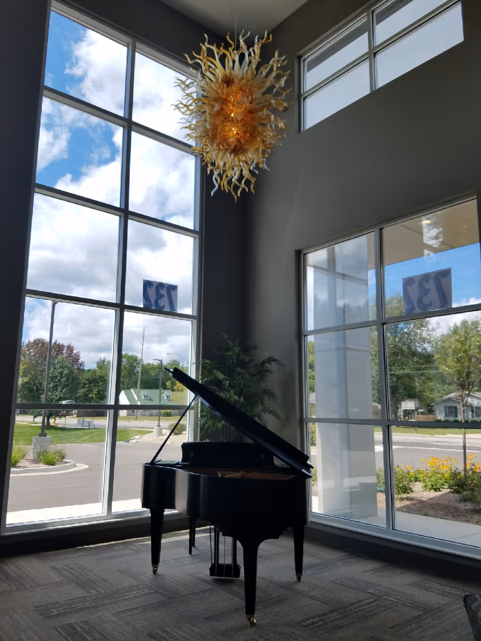 A black grand piano sits in a spacious room with large floor-to-ceiling windows letting in natural light. Outside, a street and greenery are visible. A decorative, sun-like chandelier hangs from the ceiling above the piano.