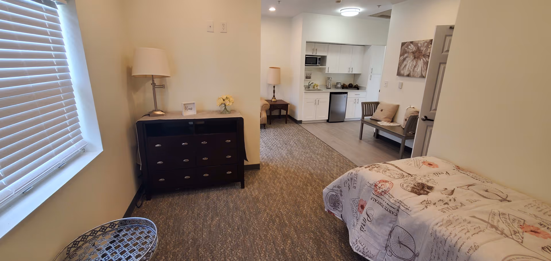 Interior view of a senior living facility room at St James Manor & Villas featuring a bed with printed bedding on the right, a dark wooden dresser with a lamp and flowers on top on the left, a window with blinds, and a small kitchenette area with white cabinets, a microwave, and a mini fridge in the background. There is also a bench with cushions and a side table with a lamp near the kitchenette.