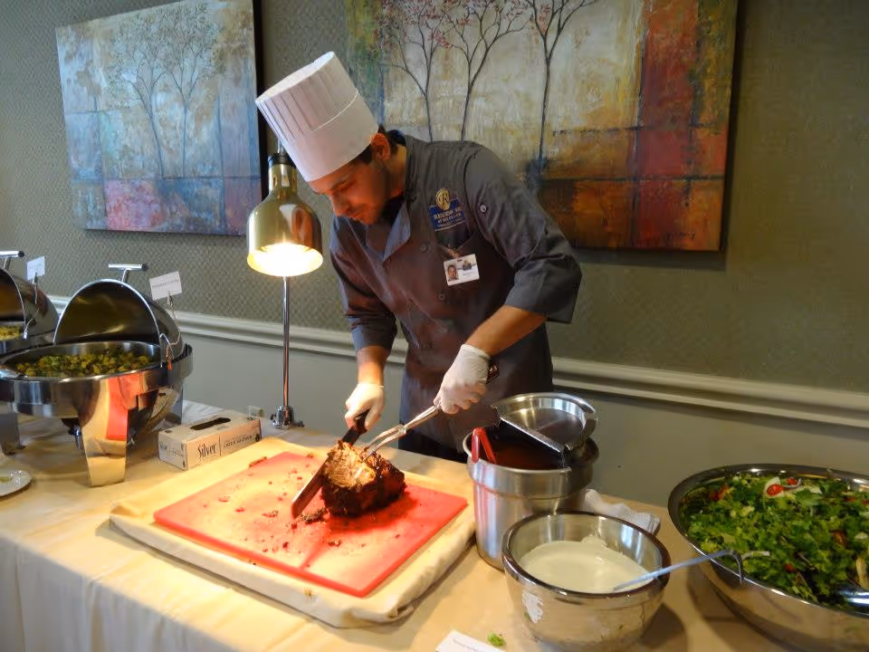 A chef wearing a white hat and gloves carves a roast at a buffet table with chafing dishes and a bowl of salad.