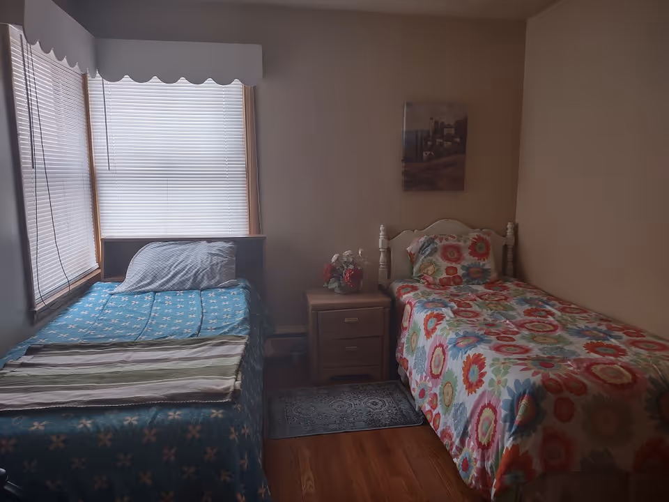 A small bedroom with two single beds separated by a wooden nightstand. The bed on the left has a blue bedspread with a floral pattern and a striped blanket at the foot. The bed on the right has a colorful floral bedspread. There is a small bouquet of flowers on the nightstand and a framed picture hanging on the beige wall above the right bed. Two windows with closed blinds are on the left side of the room.