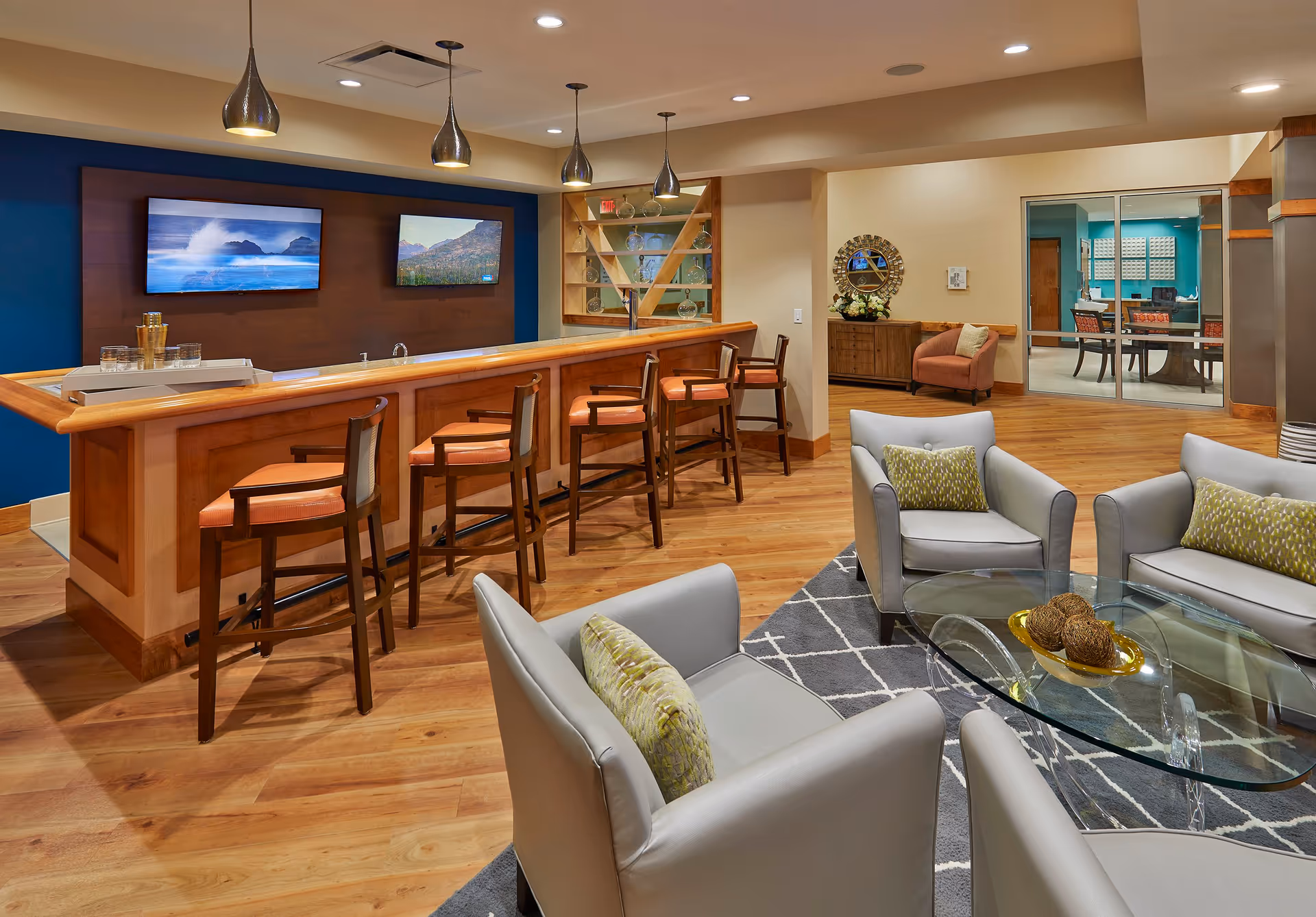 Communal lounge with a wooden bar and stools, armchairs around a glass coffee table, and wall-mounted TVs.