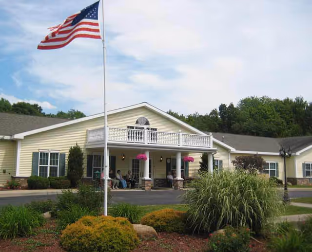 Front exterior of a light-yellow senior living building with a flagpole, covered entrance porch with people seated, and landscaped grounds.