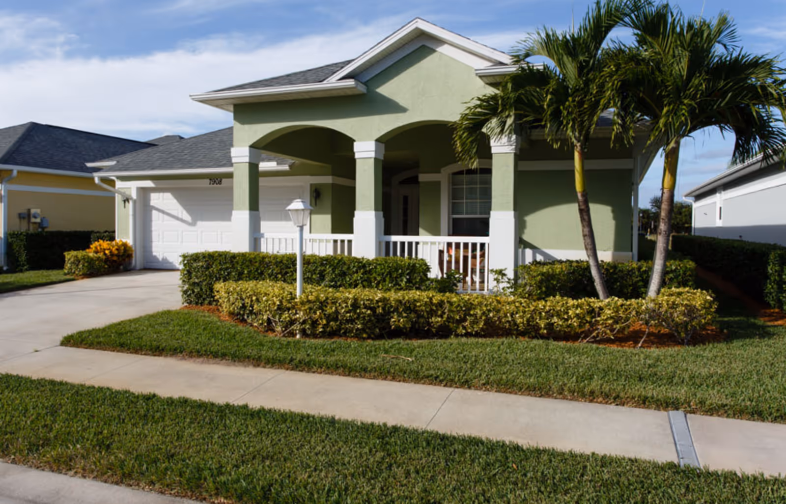 Single-story green house with a covered front porch, attached garage, manicured lawn, and palm trees.