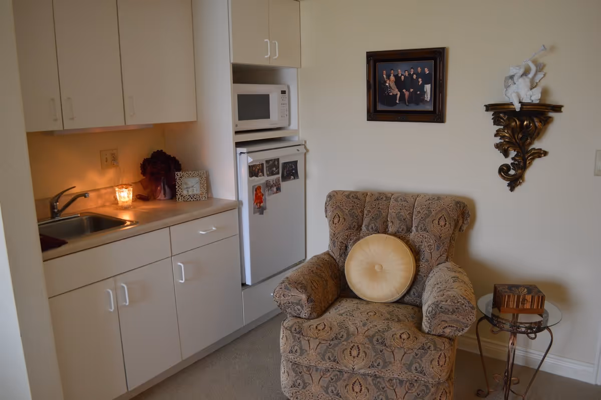A small interior room with a kitchenette and a patterned armchair topped by a round cushion, a microwave over a mini-fridge, and framed photo and decorative shelf on the wall.
