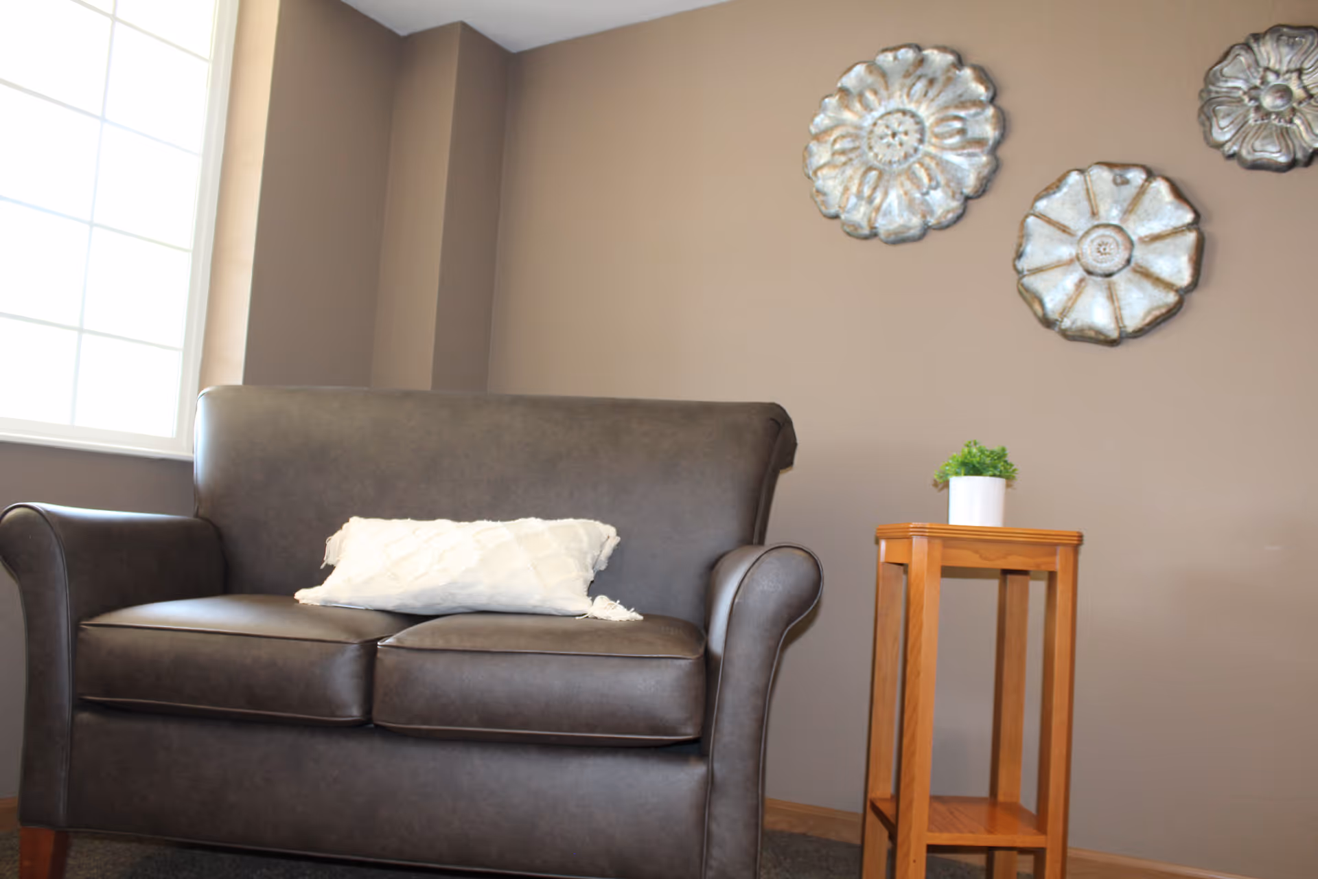 A cozy seating area with a dark brown leather loveseat featuring a white textured pillow, next to a small wooden side table with a potted green plant. The background wall is painted beige and decorated with three metallic floral wall hangings. A large window on the left allows natural light into the room.