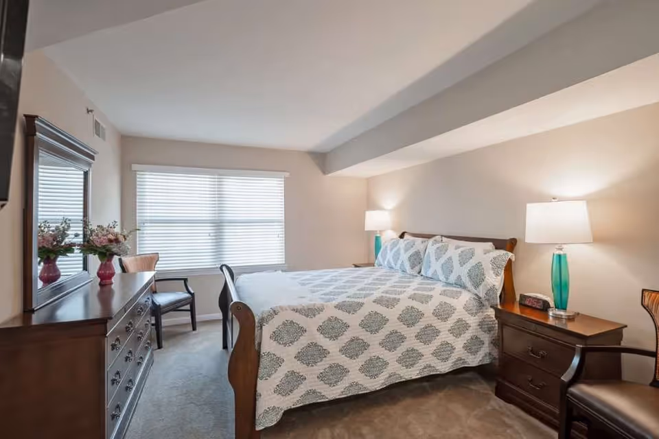 A well-lit bedroom with a large bed covered in a white and gray patterned quilt. There are two bedside tables with lamps featuring turquoise bases, a wooden dresser with a mirror, a chair, and a window with closed blinds.