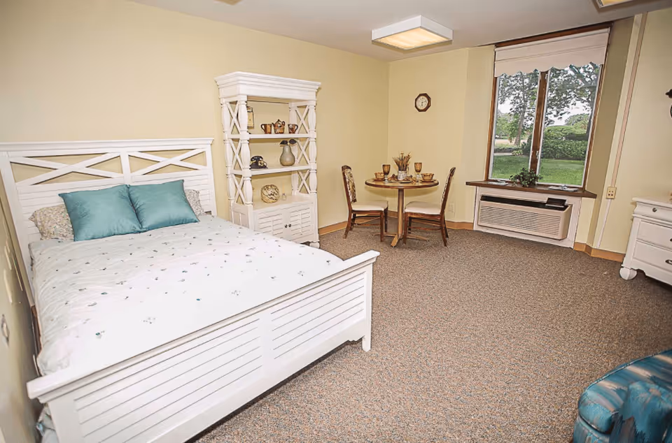 A senior living facility bedroom with a white wooden bed featuring teal pillows and a light-colored bedspread. There is a white shelving unit with decorative items, a small round dining table with two chairs set with dishes and glasses, a window showing greenery outside, and a wall-mounted air conditioning unit below the window. The room has beige walls and carpeted flooring.