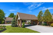 Brick senior living facility entrance with a covered porte-cochère, driveway and landscaped lawn under a blue sky.