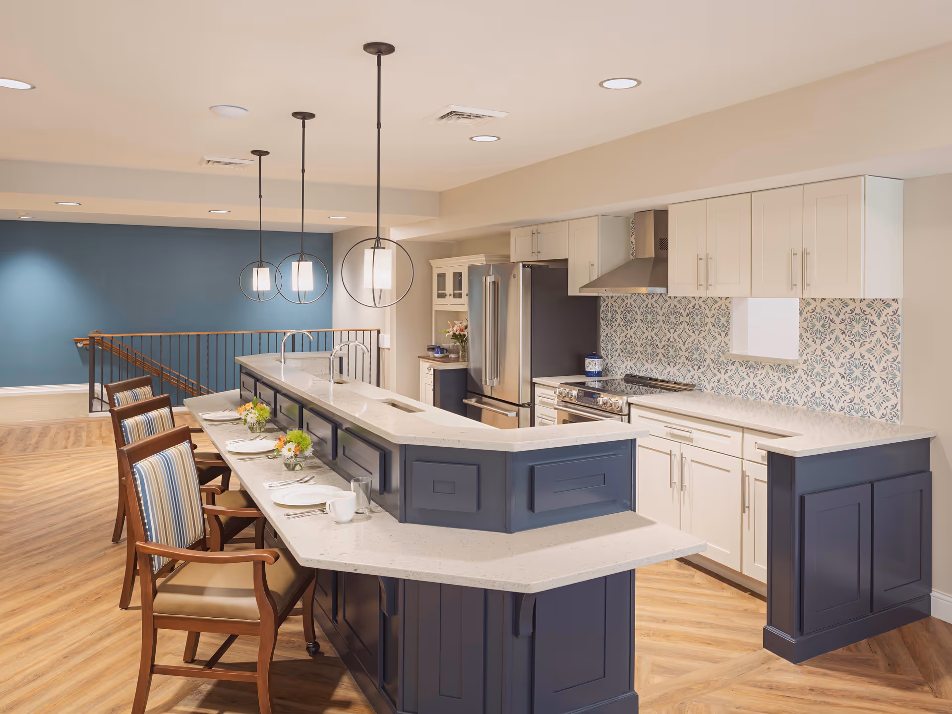 Modern kitchen area with a long island counter featuring a raised bar section and seating for four with wooden chairs. The kitchen has white cabinets, a stainless steel refrigerator, stove, and a decorative blue and white tile backsplash. Three pendant lights hang above the island, and the floor is a light wood pattern. A blue accent wall and a staircase railing are visible in the background.