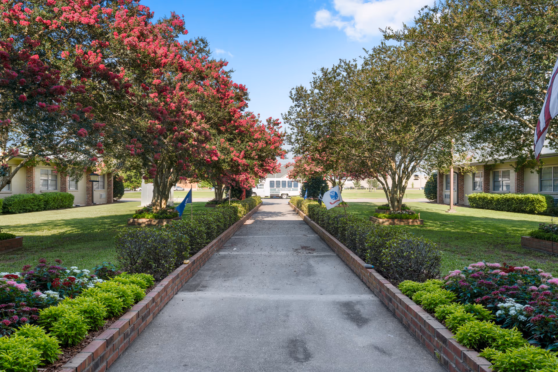 A paved walkway lined with low brick walls and green shrubs leads through a garden area with blooming trees featuring pink flowers. On either side of the walkway are well-maintained lawns and single-story buildings with windows. A white vehicle is parked at the end of the path under a blue sky with some clouds.