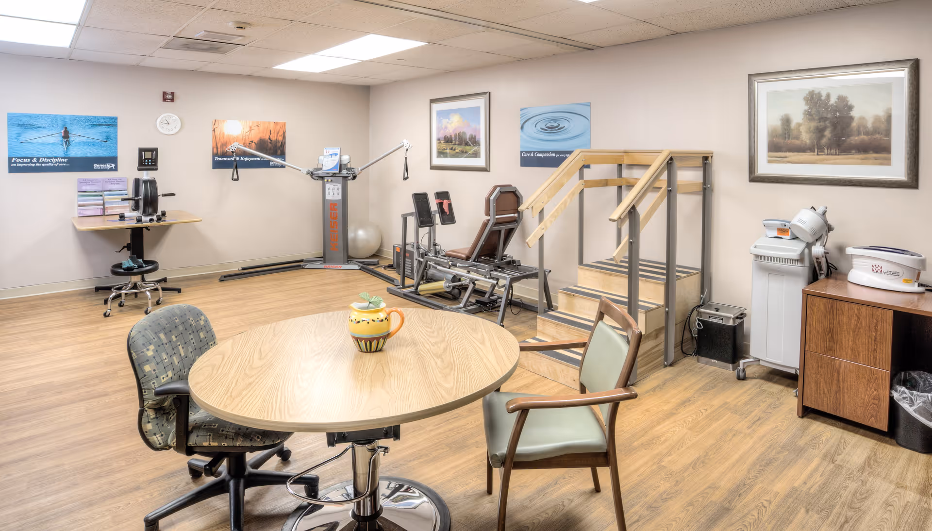 A rehabilitation therapy room with exercise equipment including a leg press machine, parallel bars with stairs for walking practice, and a therapy ball. There is a round wooden table with two chairs in the foreground, and motivational posters on the walls. The room has wood flooring and neutral-colored walls with framed artwork.