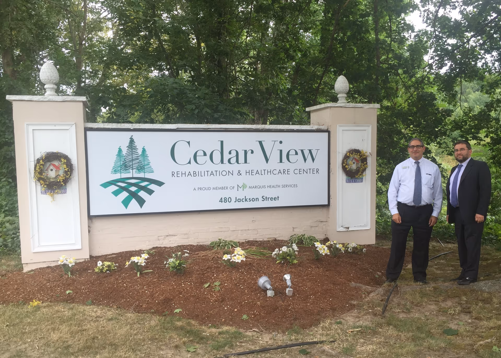 Two men in business attire standing next to a large outdoor sign for Cedar View Rehabilitation & Healthcare Center, surrounded by greenery and small flower beds.