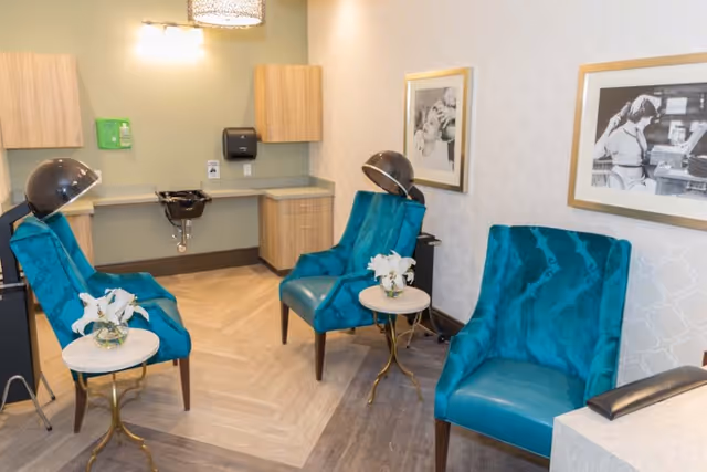 A cozy salon area with three teal salon chairs, two of which have hair dryers attached overhead. There are two small round tables with white flowers in glass vases. The room has light-colored walls with framed black and white photos, wooden cabinets, a sink, and a paper towel dispenser.