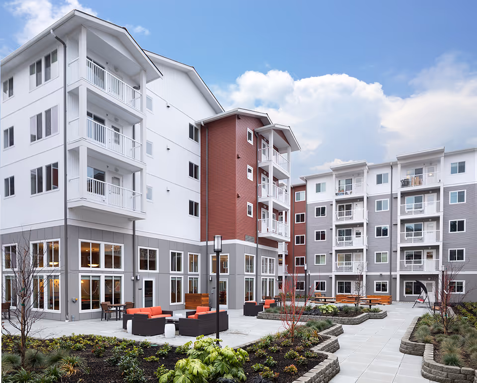 Outdoor courtyard with seating, landscaped planters and walkways surrounded by a multi-story apartment building with balconies under a blue sky.