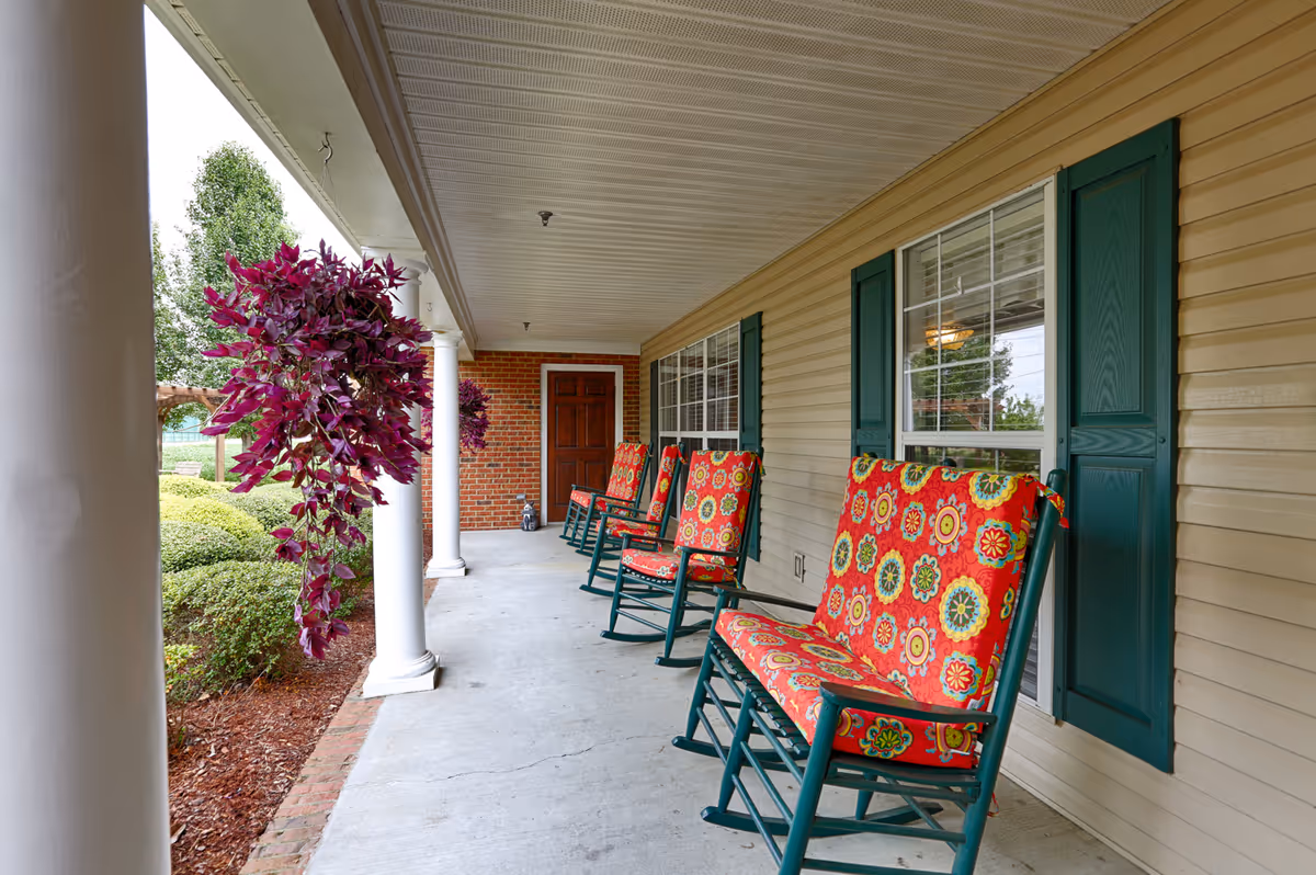 Covered front porch with white columns and four green rocking chairs with colorful red floral cushions. Hanging purple plants are visible on the left side, with green bushes and a brick wall at the far end near a wooden door.