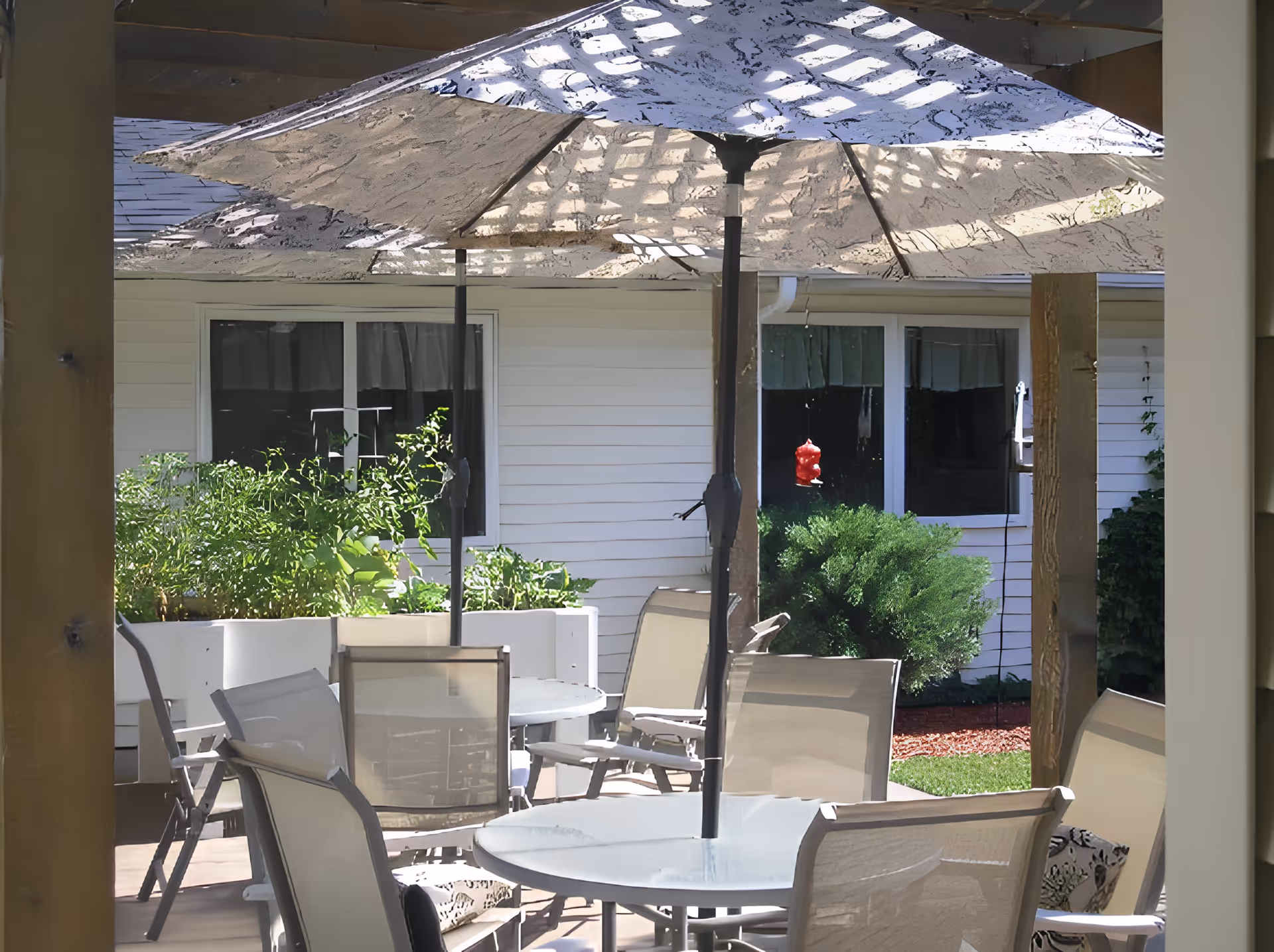 Outdoor patio area with several round glass tables and beige mesh chairs arranged around them. Large beige umbrellas provide shade over the tables. In the background, there are white siding walls with windows, green bushes, and plants in raised white planters. A red hummingbird feeder hangs near one of the windows.