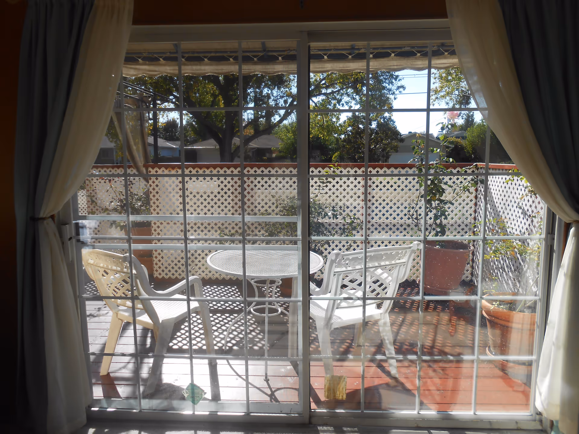 View through a glass sliding door with white curtains tied back, showing an outdoor patio area with a white metal round table, two white plastic chairs, potted plants, and a white lattice fence. Trees and houses are visible in the background under a clear sky.