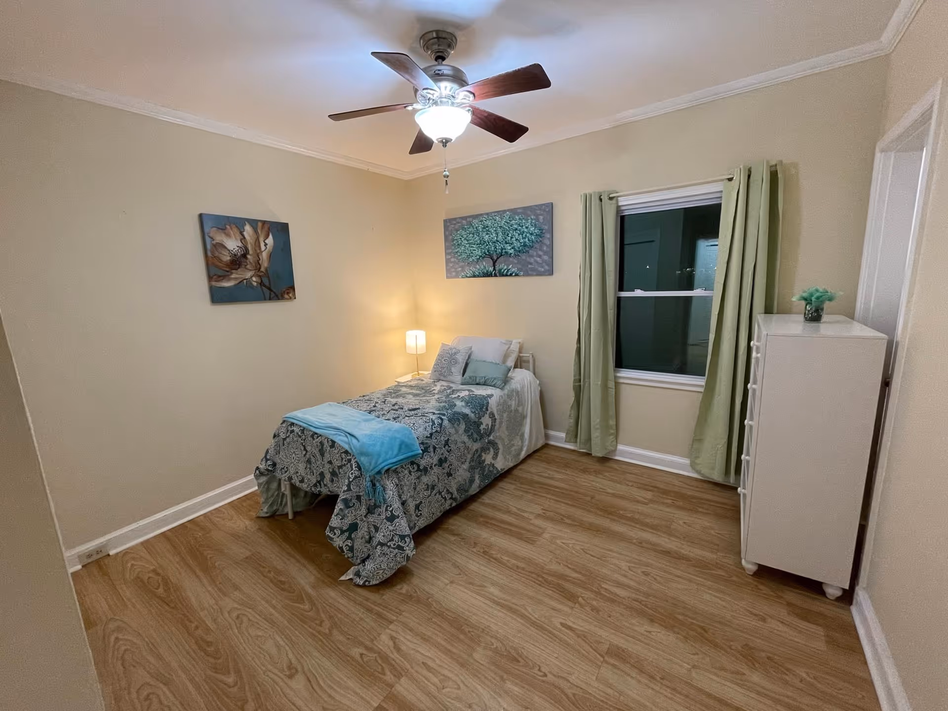 Small furnished bedroom with a single bed, ceiling fan, dresser, window with green curtains, and wall art.