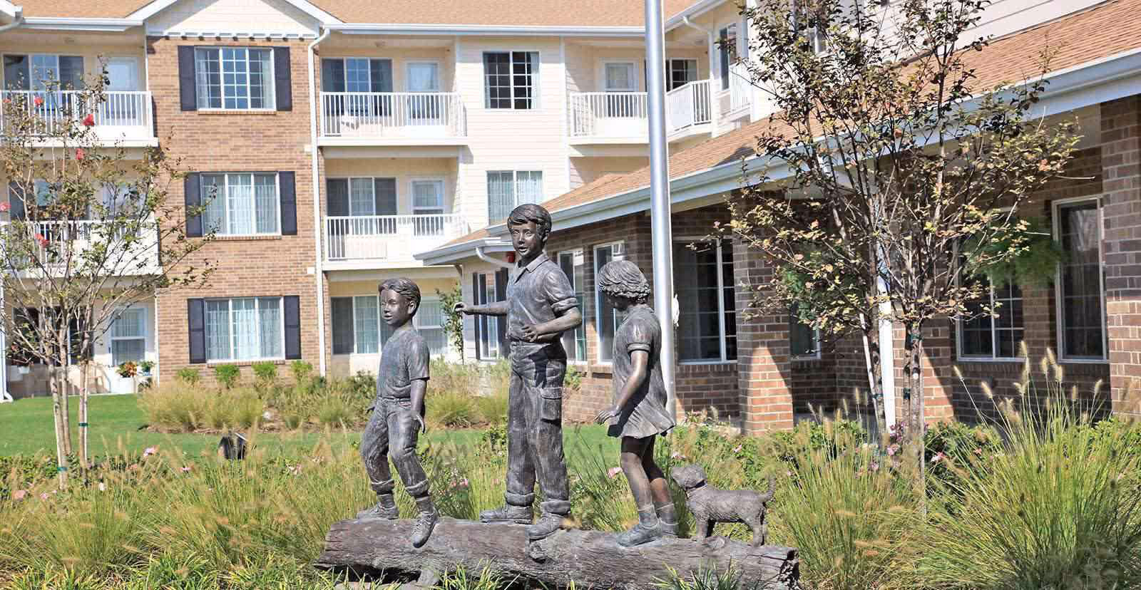 Outdoor courtyard area of a senior living facility featuring a bronze statue of three children and a dog standing on a log, surrounded by grass, trees, and flowers. The background shows a multi-story brick and siding building with balconies and windows.