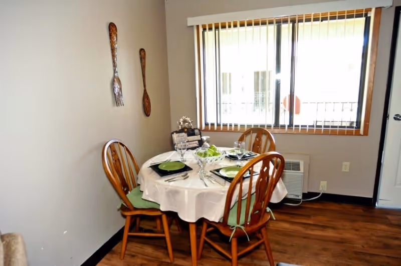A small dining area with a round table covered with a white tablecloth set for four people. The table has green plates, glasses, and silverware. Four wooden chairs with green cushions surround the table. On the wall, there are large decorative wooden spoon and fork hanging. A window with vertical blinds is in the background, and a small air conditioning unit is below the window.