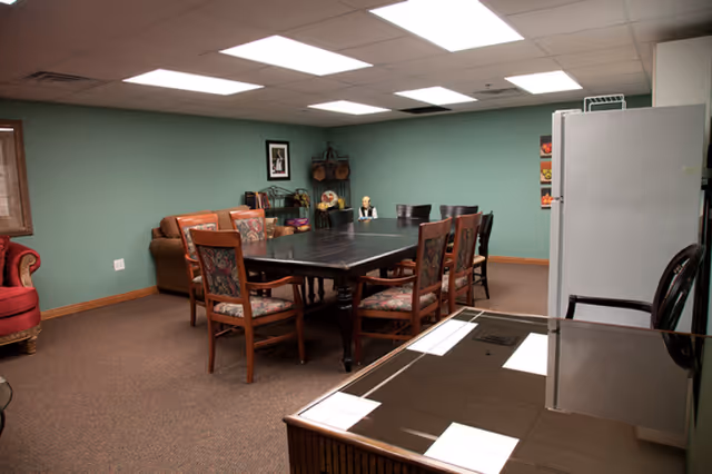 A carpeted communal room with a large dark dining table surrounded by chairs, a refrigerator, couch and armchair against teal walls.
