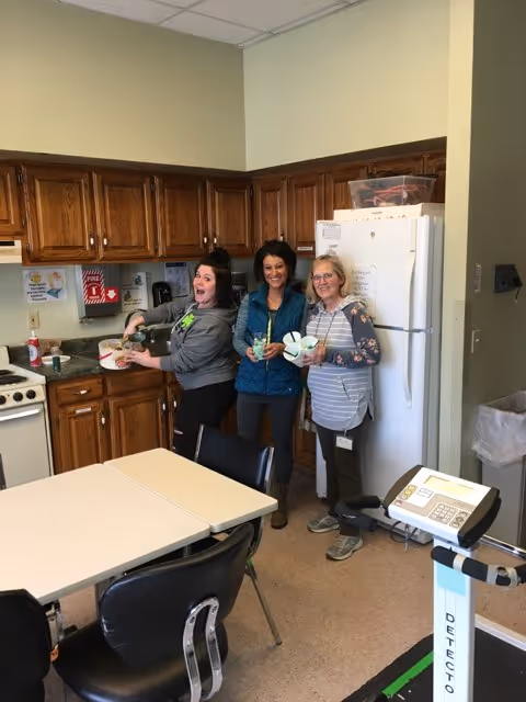 Three people smiling and holding bowls in a communal kitchen with wooden cabinets, a white refrigerator, and a table.