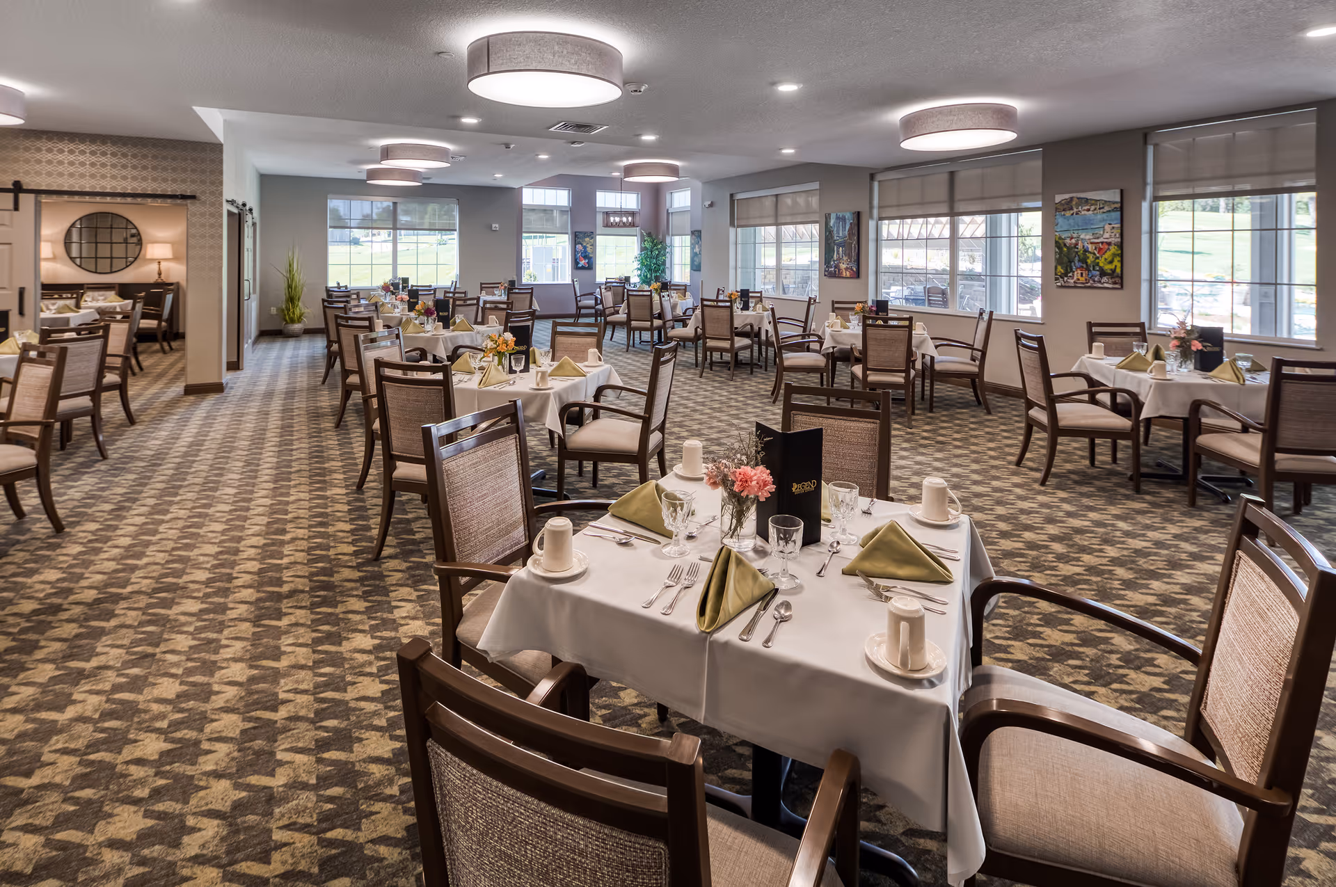 A spacious dining room with multiple tables covered in white tablecloths, each set with green folded napkins, cups, glasses, and silverware. The room has large windows letting in natural light, patterned carpet flooring, and modern ceiling lights. There are paintings on the walls and a small plant near the windows.