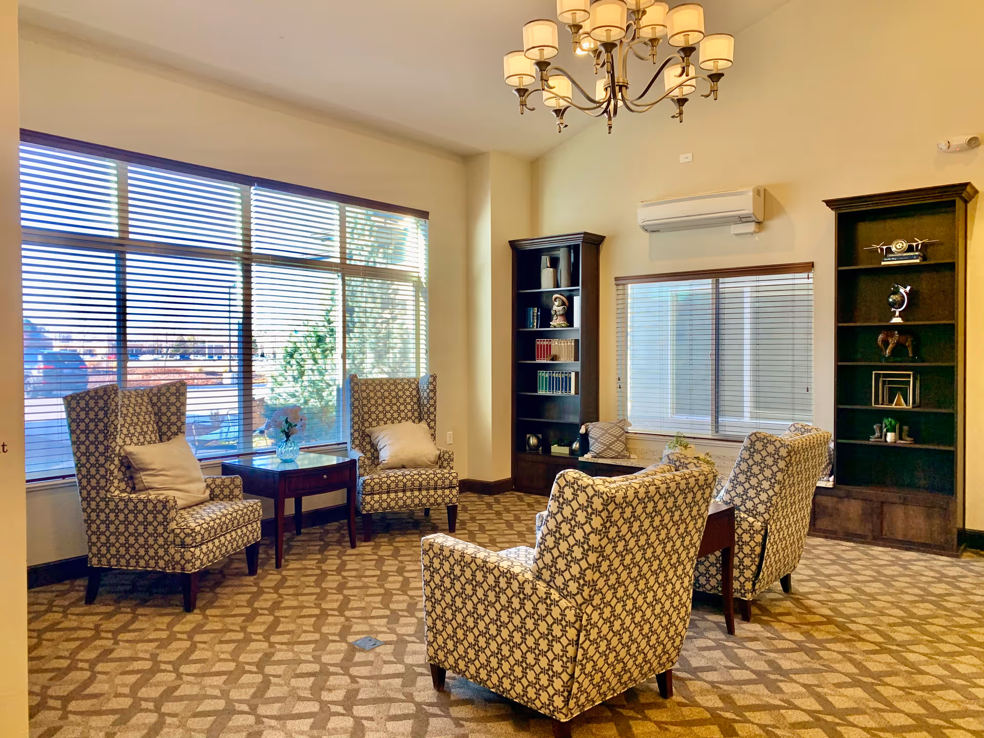 A bright and cozy living room area with four patterned armchairs arranged around a wooden coffee table. The room features large windows with blinds, two tall dark wood bookshelves filled with decorative items and books, and a chandelier hanging from the ceiling. The carpet has a geometric pattern and the walls are painted a light color.