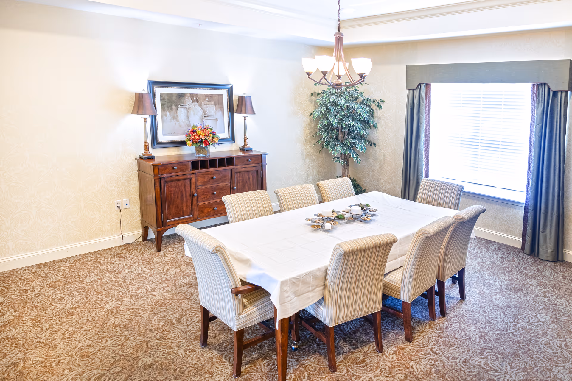 A dining room with a rectangular table covered with a white tablecloth surrounded by eight striped upholstered chairs. A wooden sideboard with two lamps and a framed painting above it is against the wall. A large window with curtains lets in natural light, and a chandelier hangs from the ceiling. There is a decorative plant in the corner and patterned carpet on the floor.