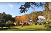 A large green lawn with trees showing autumn foliage in the background and a white multi-story building partially visible behind the trees under a blue sky with a few clouds.