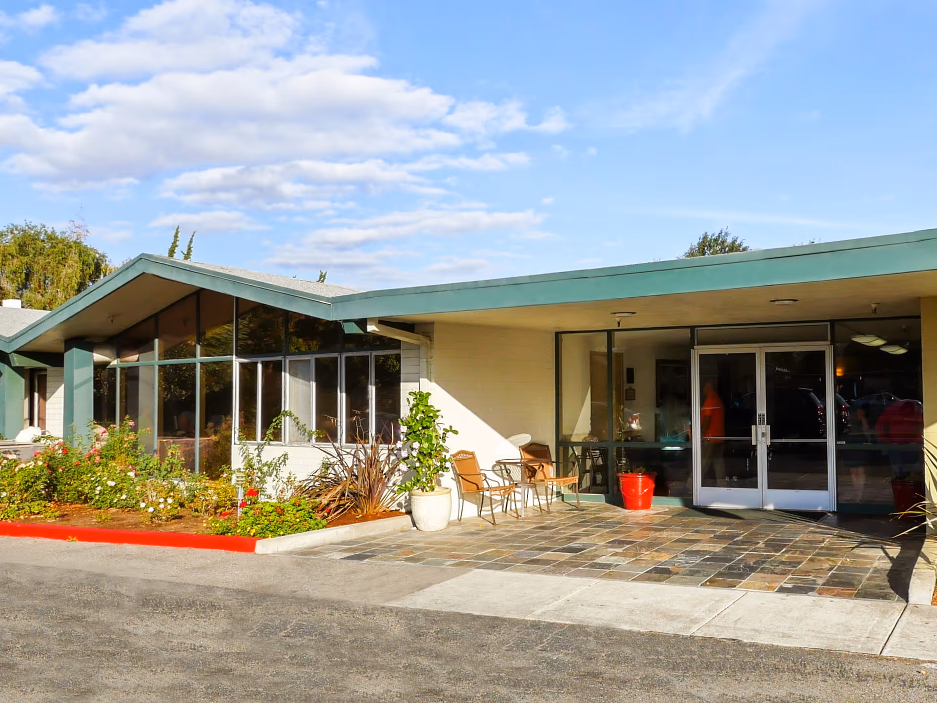 Exterior view of the entrance to Grant Cuesta Sub-acute and Rehabilitation Center, featuring large glass windows, a covered entryway with double glass doors, two chairs with a small table, potted plants, and a tiled walkway under a partly cloudy sky.