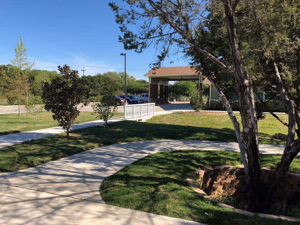 Outdoor view of a senior living facility with a curved concrete walkway, green grass, several trees, and a covered entrance area with parked cars in the background under a clear blue sky.