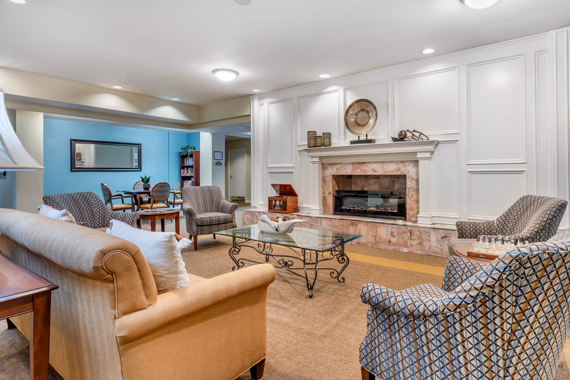 A cozy living room area in a senior living facility featuring a beige sofa with white pillows, patterned armchairs, a glass coffee table with decorative items, and a fireplace with a marble surround and white paneled wall. In the background, there is a small dining area with a round table and chairs, a bookshelf, and a large mirror on a blue accent wall.