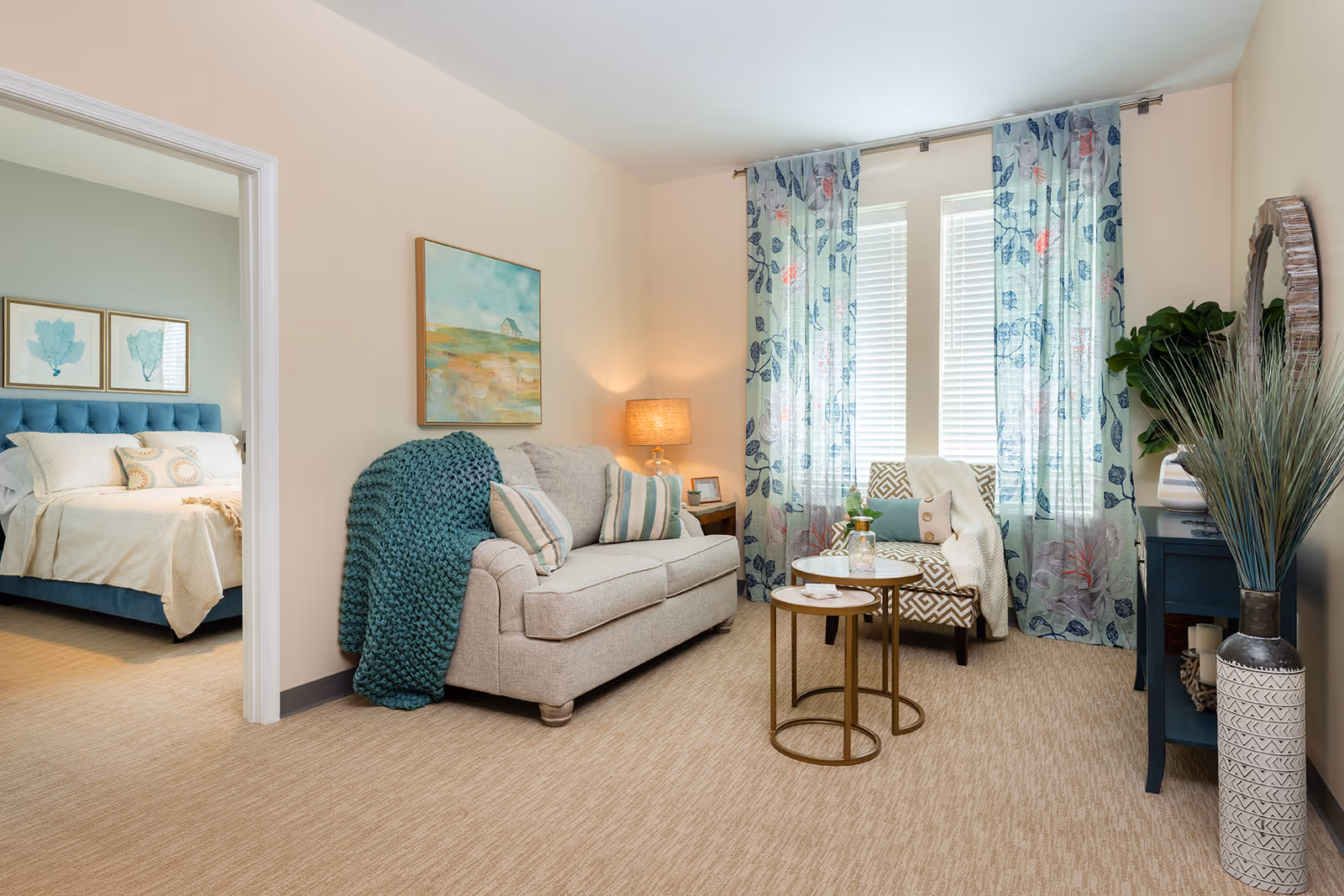 A cozy living room with a beige sofa adorned with striped pillows and a teal knitted throw blanket. Next to the sofa is a small side table with a lamp and picture frame. Two round nesting coffee tables with a small plant and glass pitcher are in front of the sofa. A patterned armchair with a white throw blanket is positioned near two tall windows with floral curtains. A blue console table with decorative items and a large vase with tall grass-like plants is against the wall. Through an open doorway, a bedroom with a blue upholstered headboard and framed artwork above the bed is visible.