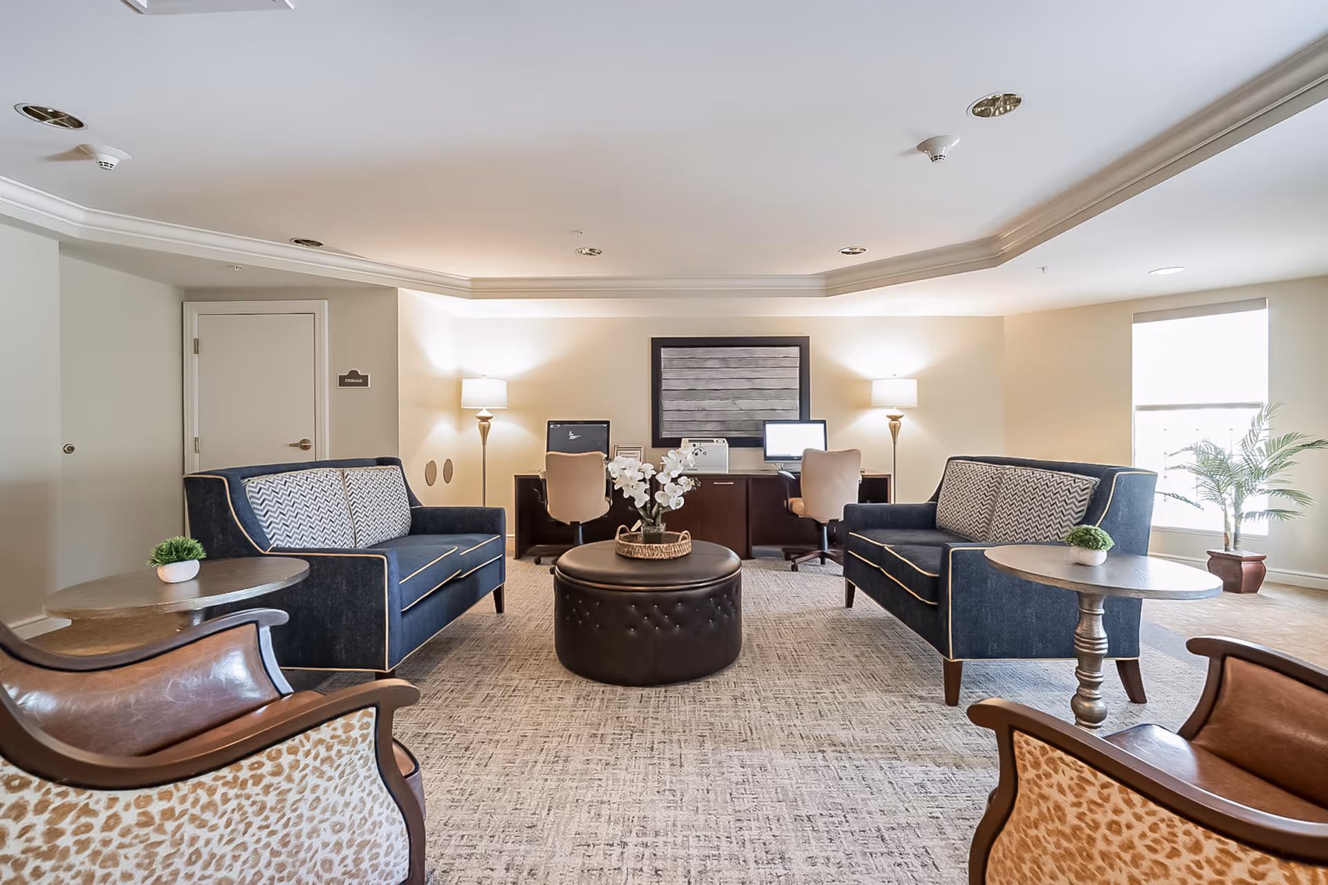 A well-lit lounge area with two blue sofas facing each other, a round black ottoman with a flower arrangement in the center, two wooden side tables with small plants, and two beige office chairs at desks with computer monitors against the back wall. The room has beige walls, a large window with natural light, and a potted plant near the window.
