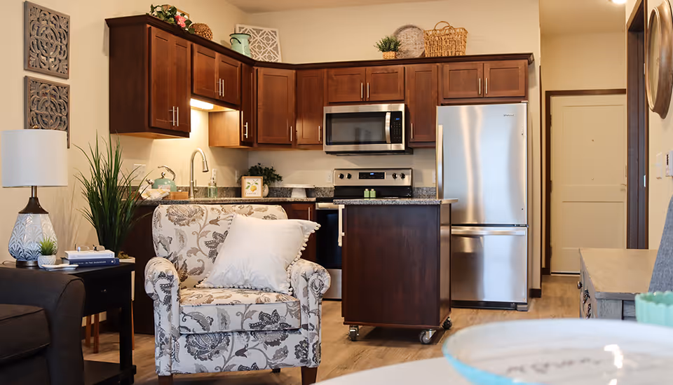 Open-plan senior apartment interior showing a small kitchen with dark wood cabinets, stainless steel appliances, a rolling island, and a patterned armchair in the foreground.