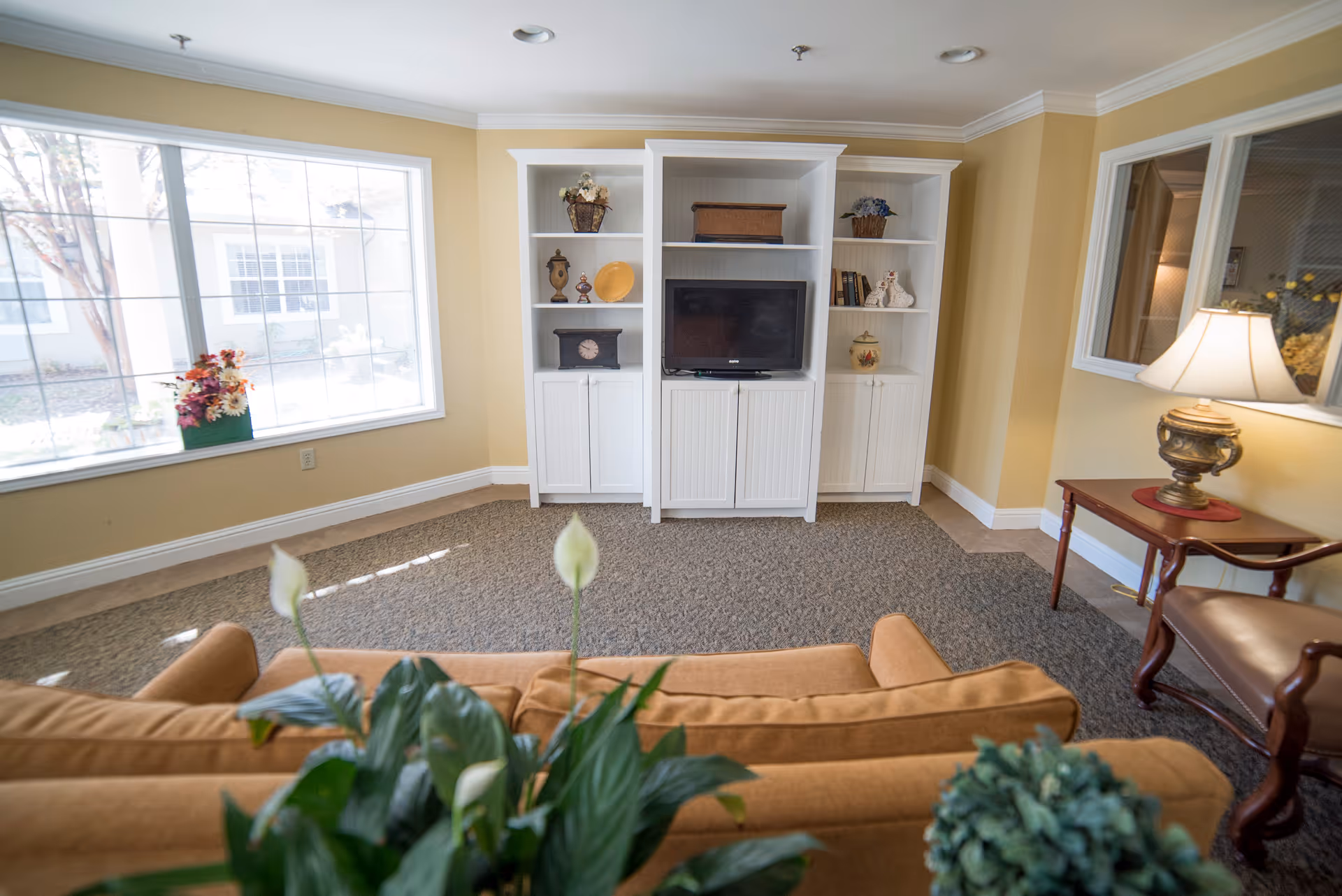 A cozy living room with a large window letting in natural light, a white entertainment center with a TV and decorative items, a brown sofa in the foreground, a side table with a lamp, and a chair. The walls are painted yellow and the floor is carpeted.