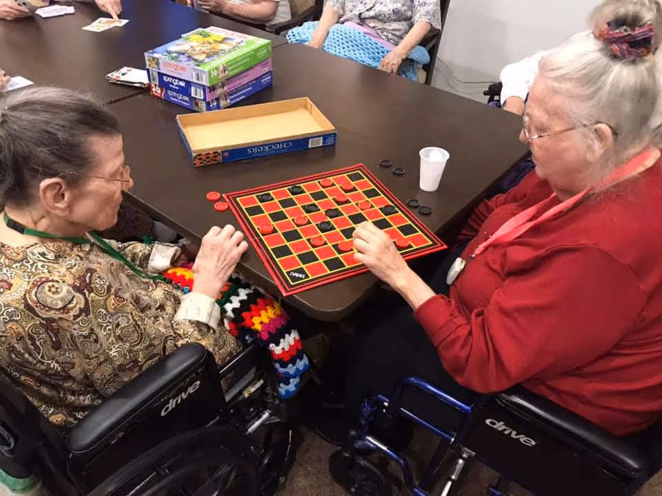 Two elderly women in wheelchairs playing a game of checkers at a table with other seniors sitting around it. There are several board game boxes on the table.