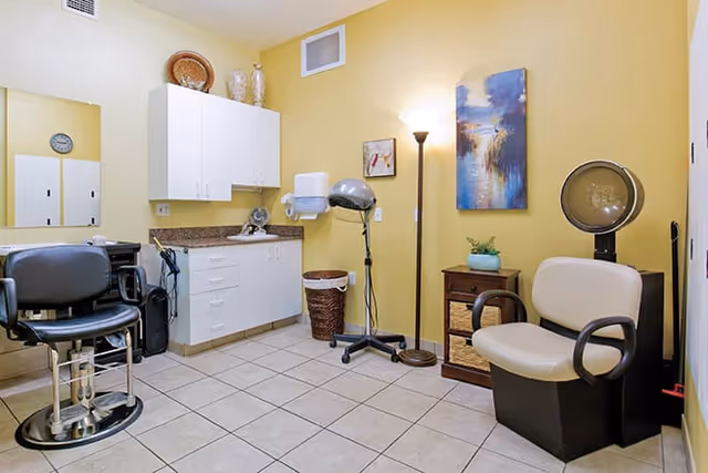 A small salon room with yellow walls and tiled floor, featuring two salon chairs, a hair dryer, a floor lamp, a small cabinet with a plant, and white cabinets mounted on the wall above a countertop.