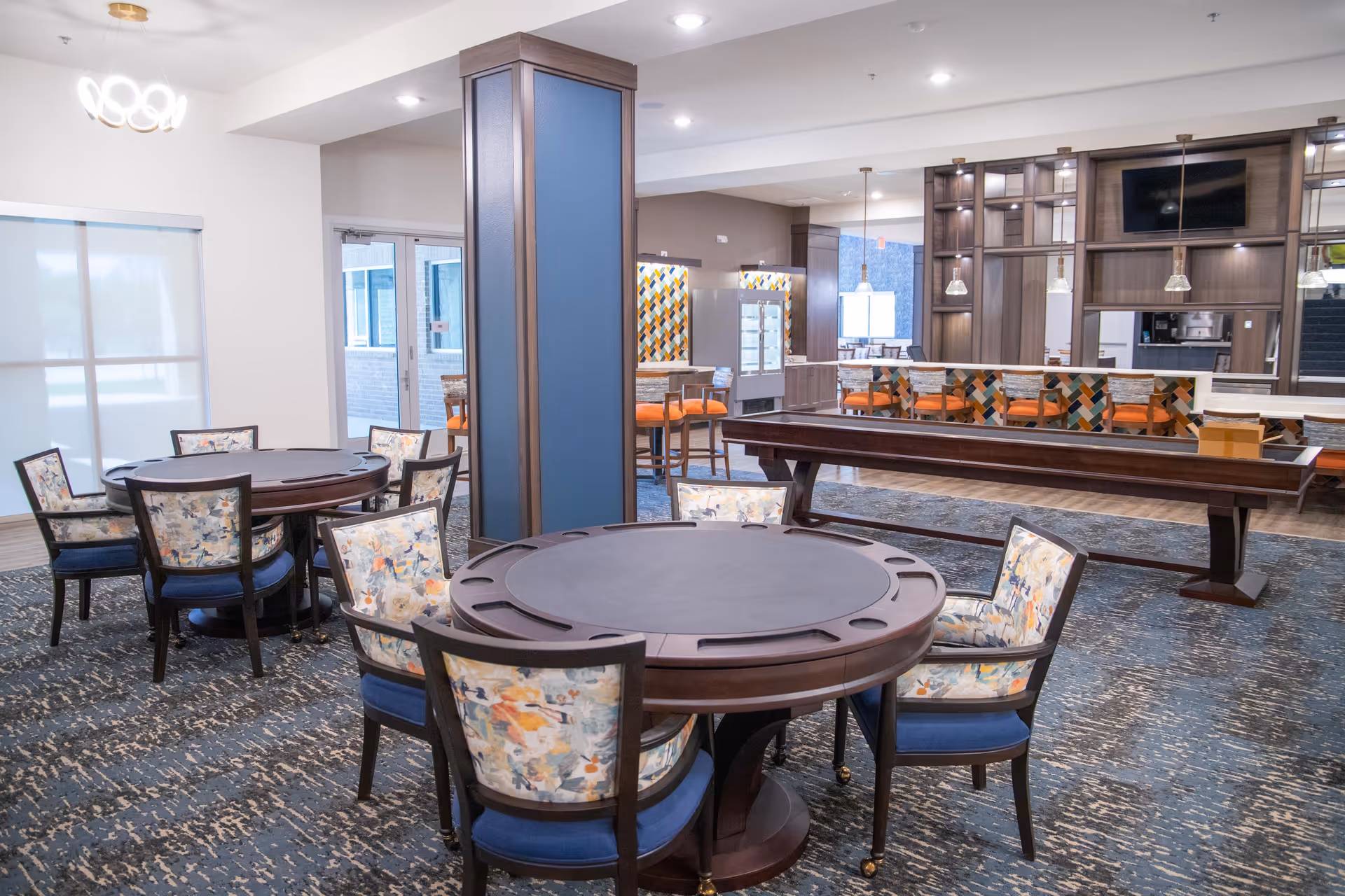 A spacious common area in a senior living facility featuring round tables with floral upholstered chairs, a pool table, and a bar-style counter with orange cushioned chairs. The room has modern lighting, carpeted flooring, and large windows with blinds.