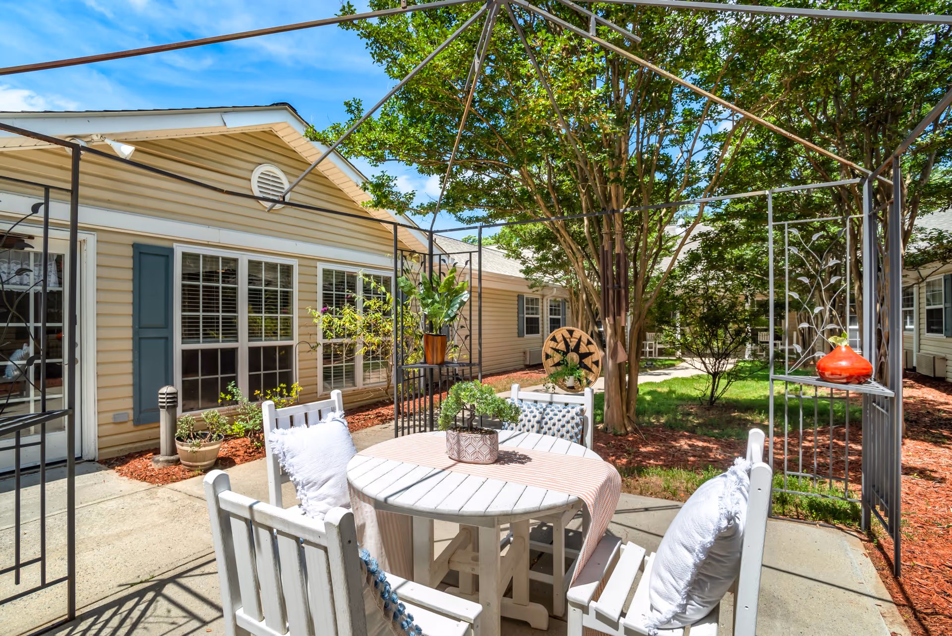 Outdoor patio area with a white round table and four white wooden chairs with cushions. The table has a small potted plant and a striped table runner. The patio is surrounded by a metal frame structure and is adjacent to a beige building with blue shutters and multiple windows. There are trees and landscaped areas with mulch and plants around the patio under a bright blue sky.