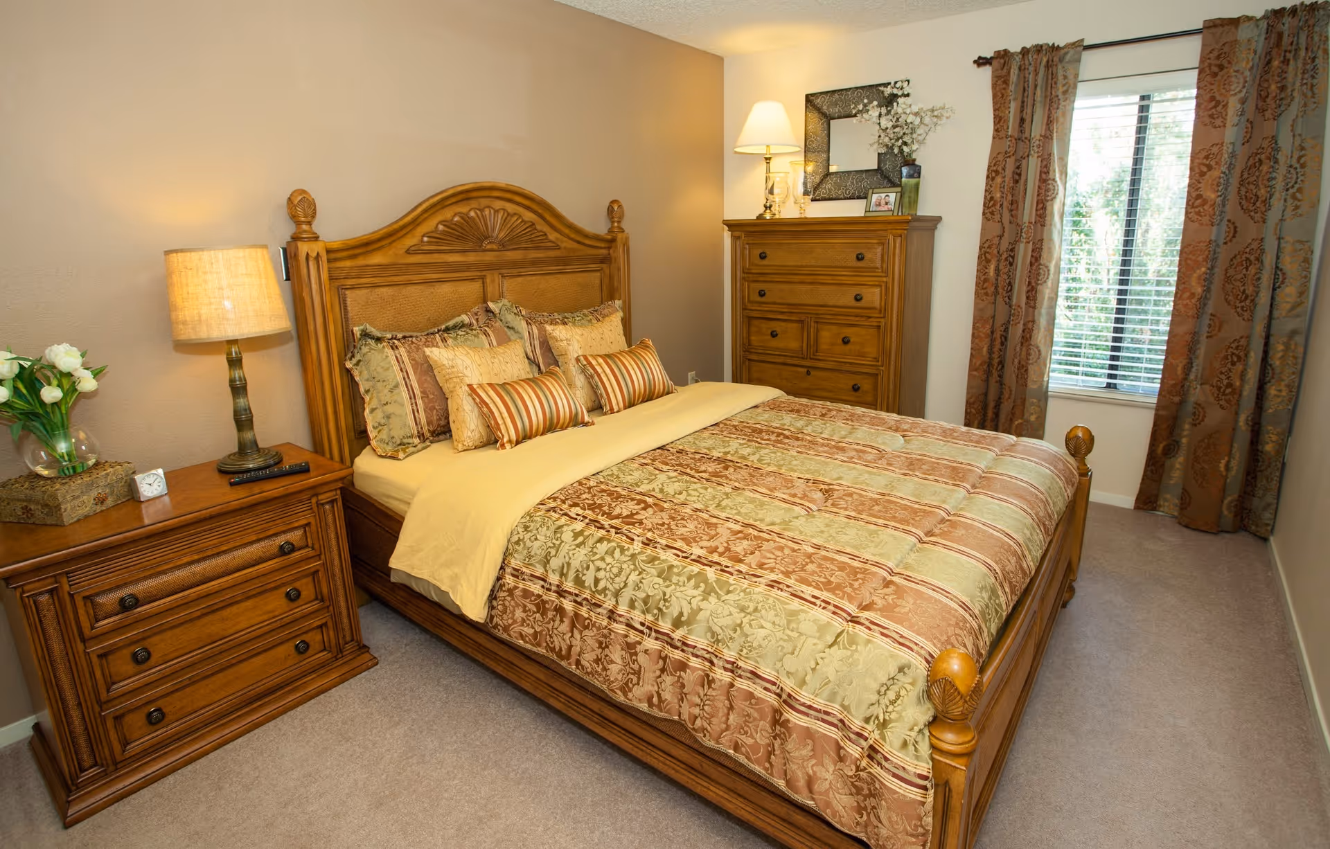 Furnished bedroom with a wooden bed dressed in patterned bedding, matching nightstand and dresser, lamp and a window with curtains.