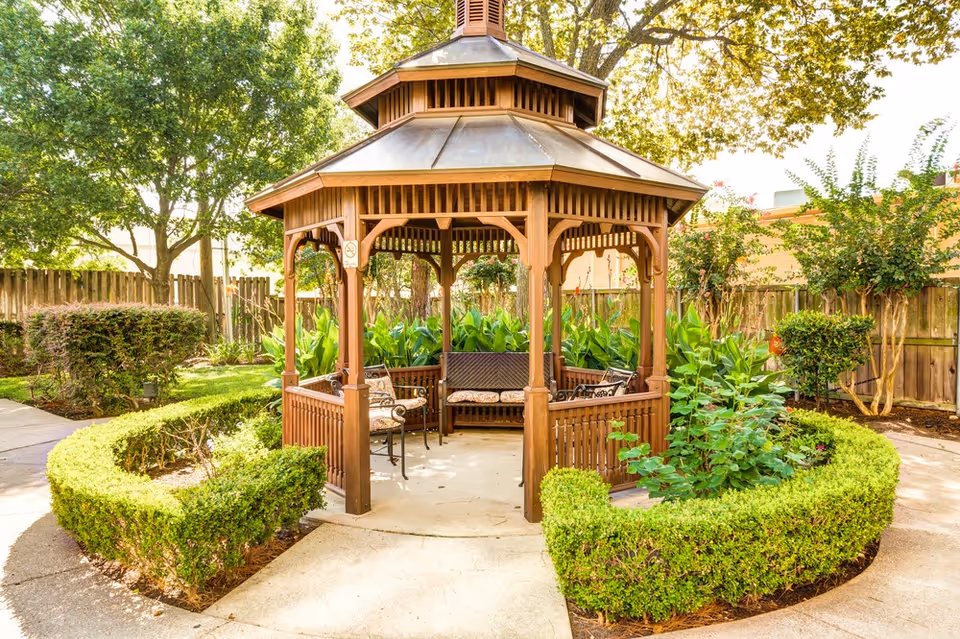 Wooden octagonal gazebo with benches and chairs surrounded by manicured hedges, plants, and trees in a sunny courtyard.