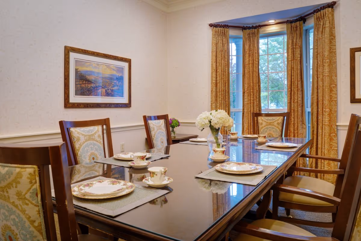 A formal dining room with a long glass-topped table set with china, wooden chairs, a vase of flowers, and bay windows with gold curtains.