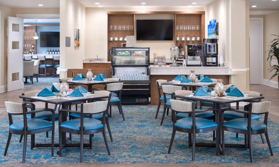 Bright dining room with tables set with teal napkins and chairs facing a buffet counter with a refrigerated display and coffee machines.