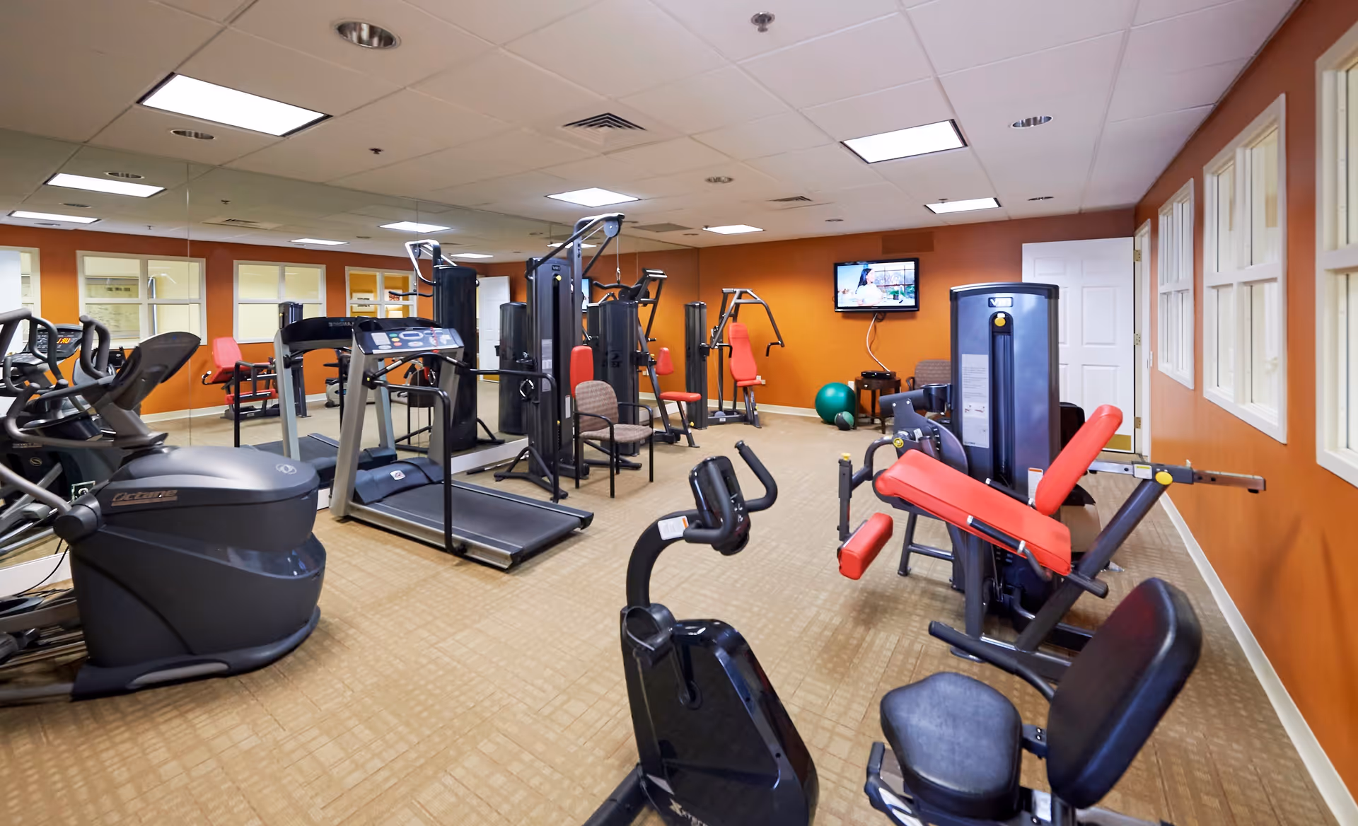 Interior view of a fitness room in a senior living facility with various exercise equipment including treadmills, stationary bikes, weight machines, and chairs. The walls are painted orange and there is a TV mounted on the far wall.