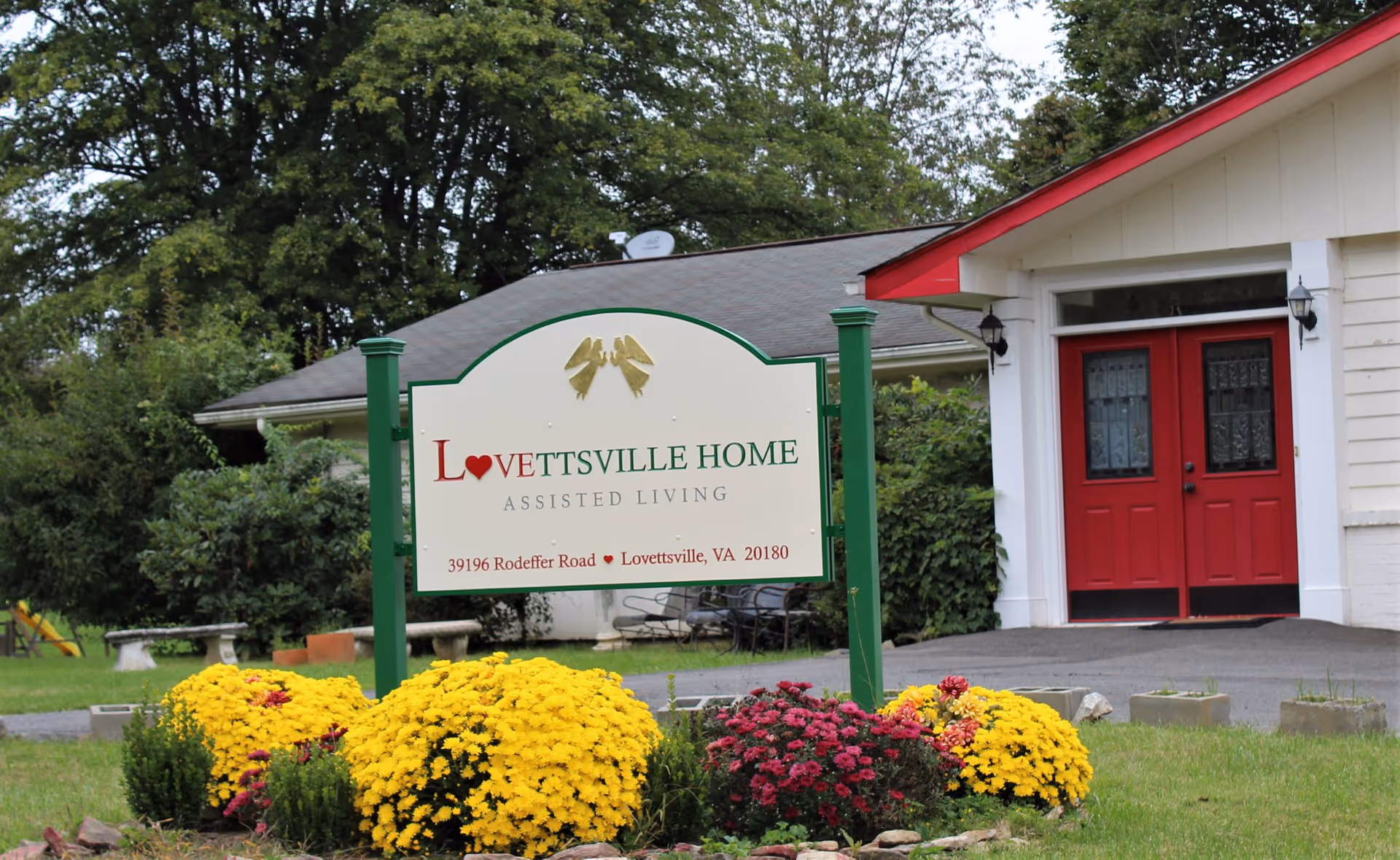 Outdoor view of Lovettsville Home Assisted Living facility sign with yellow and red flowers in front. The building with red double doors and white siding is visible in the background surrounded by greenery and trees.